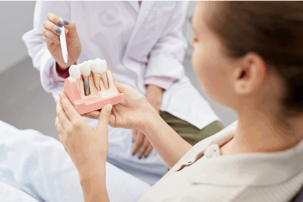 Dentist showing a patient a model of a dental implant and crown, representing a consultation on the various **Tooth Replacement Options** available at Manassra Dental in San Jose.