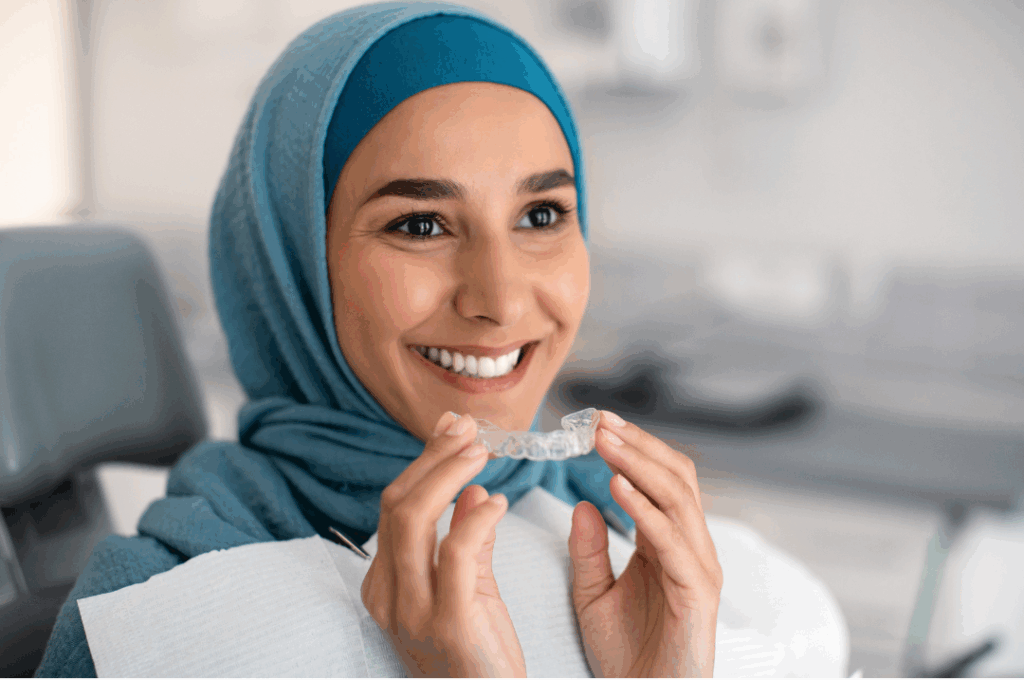 Smiling adult woman in a hijab holding a **clear Invisalign aligner** tray, highlighting discreet teeth straightening for **Adults and Teens** at Manassra Dental in San Jose.