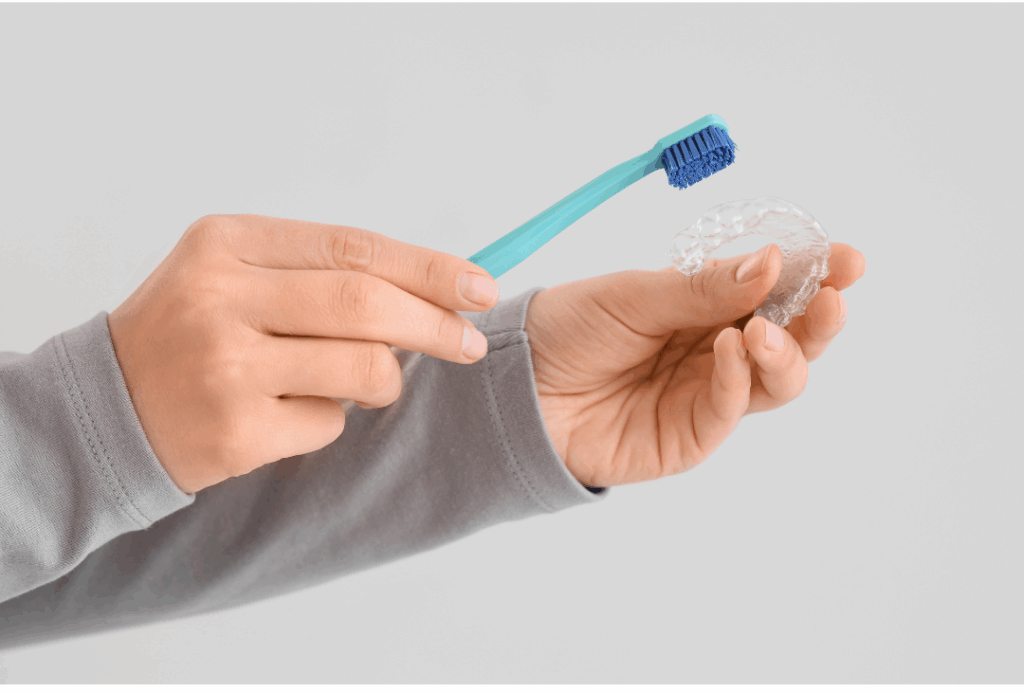 Hands holding a toothbrush and a clear dental aligner tray, demonstrating proper cleaning and **caring for aligners** to maintain hygiene at Manassra Dental in San Jose.