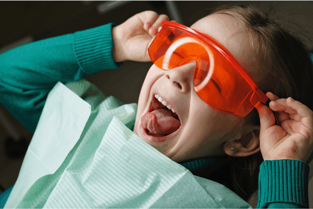Happy young girl wearing safety glasses in a dental chair, illustrating why **pediatric dentistry is different**—it's specialized, fun, and focused on children's unique developmental needs at Manassra Dental in San Jose.