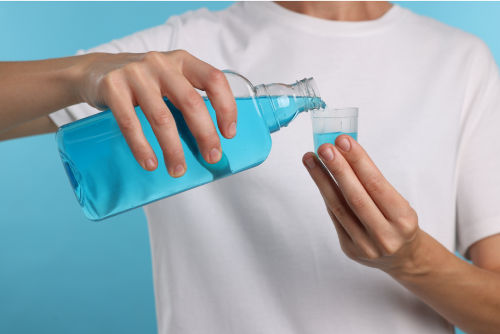 A person in a white t-shirt pouring blue fluoride mouthwash into a small plastic cup.