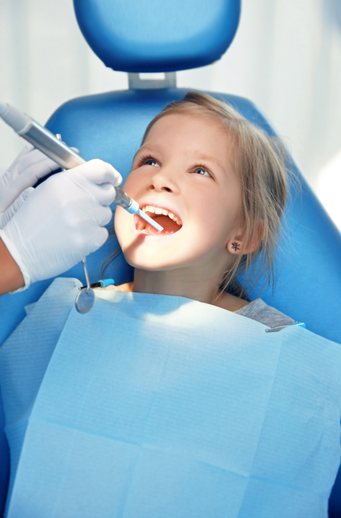 A young girl smiling with her mouth open while a dental professional applies a preventative dental sealant.