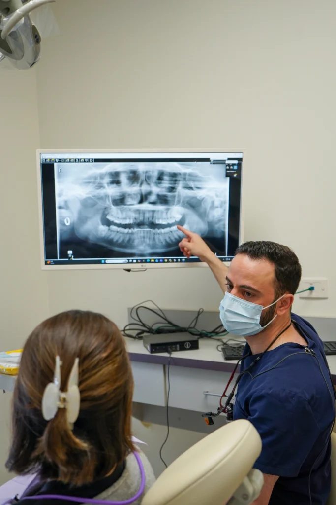 A dentist wearing a face mask pointing out specific structures on a digital dental panoramic radiograph monitor for a patient.